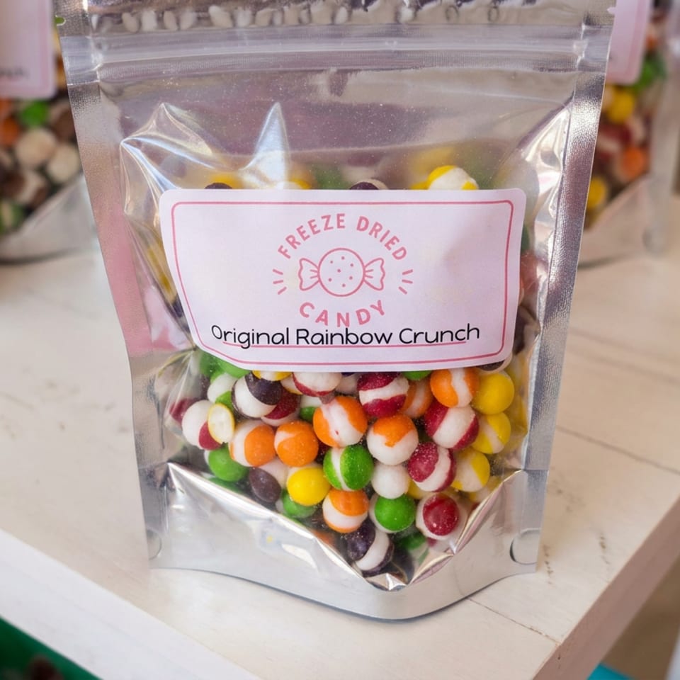 Silver pouch of freeze-dried candy labeled Original Rainbow Crunch, filled with colorful round candies on a display table.